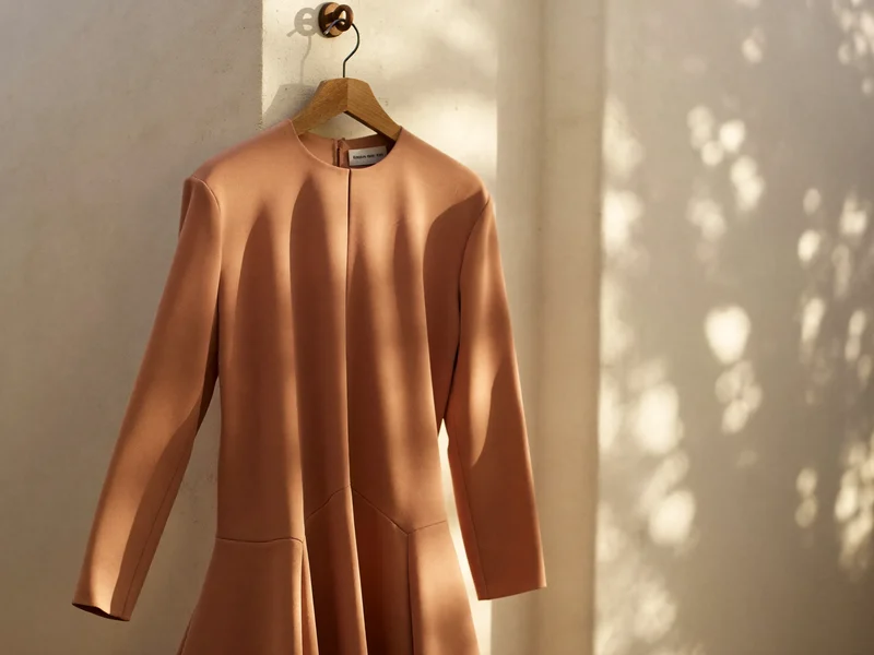 Midikleid in lehmfarbenem Stoff auf Holzbügel vor schlichter Putzwand